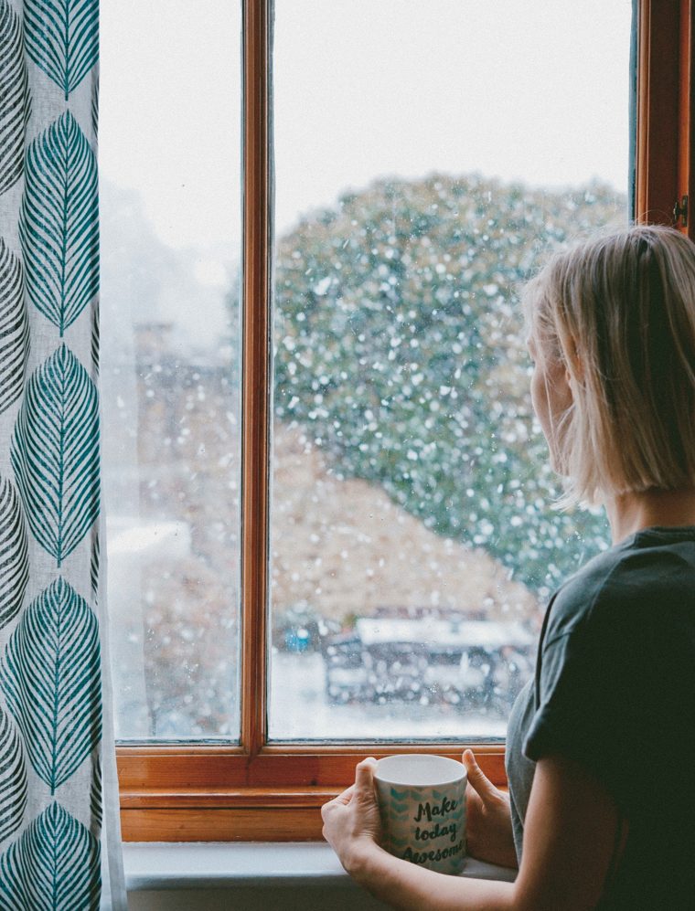 A woman holding a mug, looking out a window at falling snow