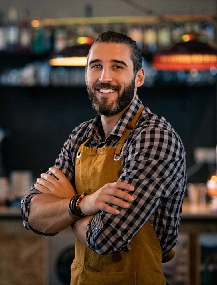 A smiling business owner standing with arms crossed in front of the bar of his café